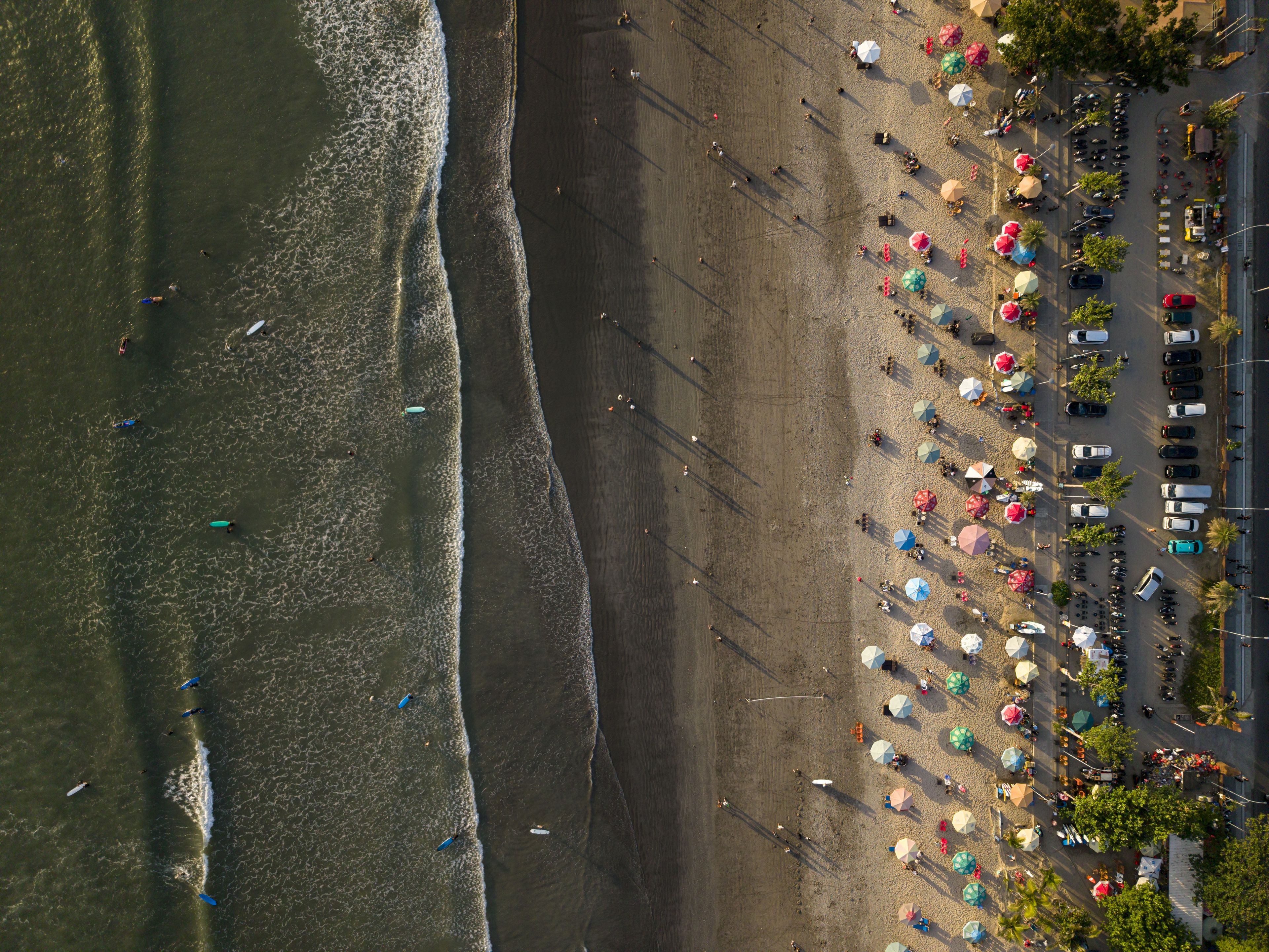 Aerial view of Kuta Beach with colorful umbrellas, people relaxing on the sand, ocean waves, and parked vehicles along the coastline in Kuta, Bali, Indonesia.