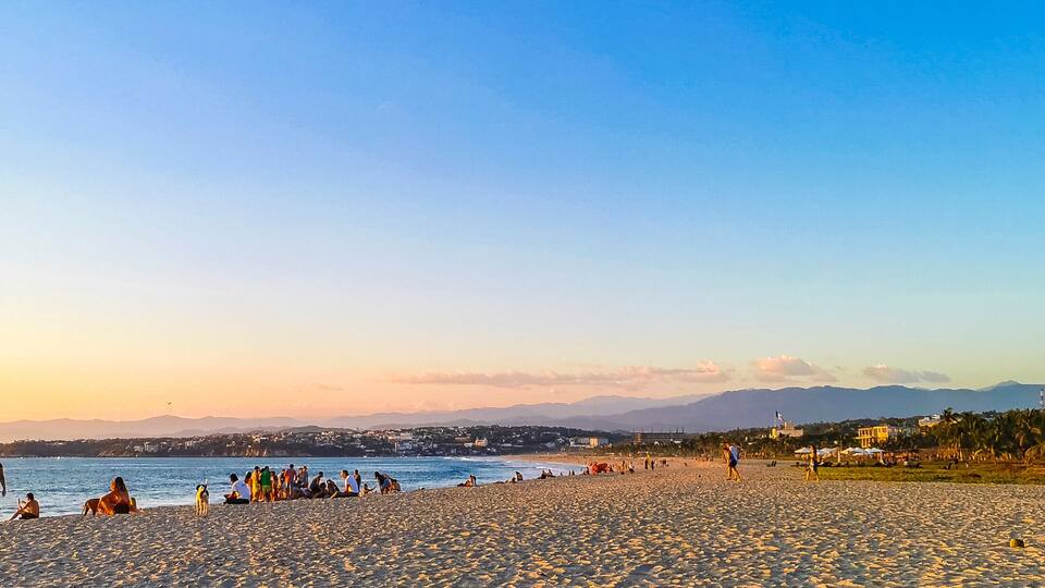 A beach with people relaxing and walking along the shore, palm trees, and mountains in the background.