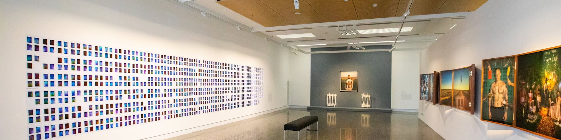 An art gallery with a large grid of colorful photographs on the left wall, a bench in the center, and paintings on the right wall.
