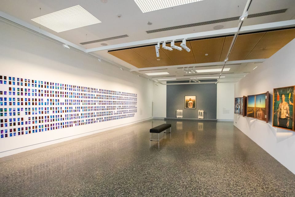 An art gallery with a large grid of colorful photographs on the left wall, a bench in the center, and paintings on the right wall.
