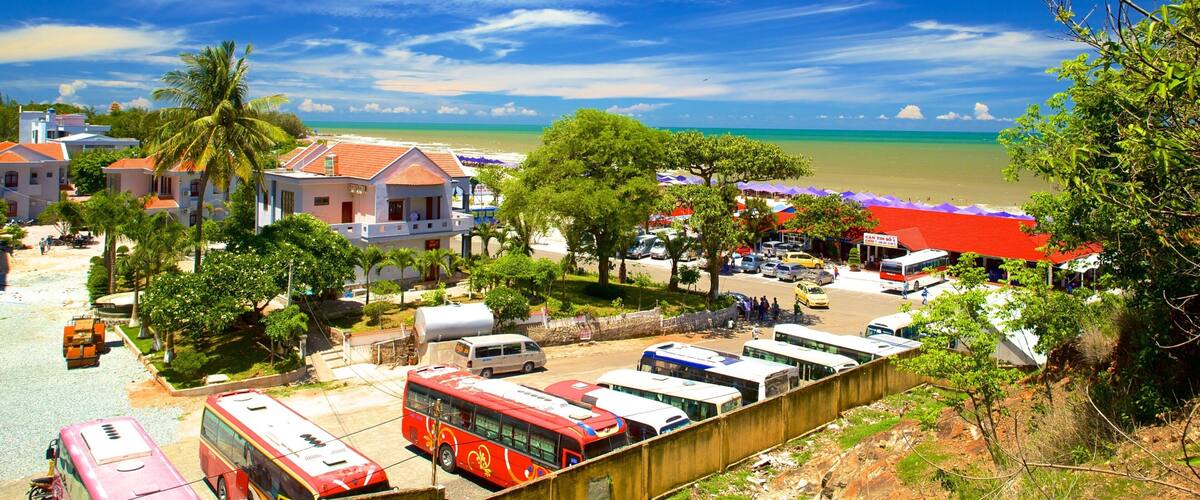A coastal town with a beach, parked buses, and palm trees.