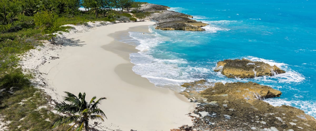 A sandy beach with turquoise waters, rocky outcrops, and palm trees.