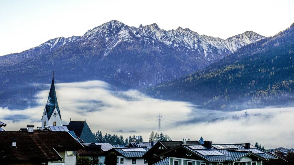 A village with traditional houses, a church with a spire, and snow-capped mountains in the background.