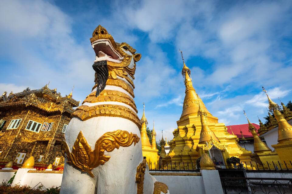 Eine goldene Statue mit Löwenkopf, ein Tempel mit rotem Dach und goldene Türme vor blauem Himmel.