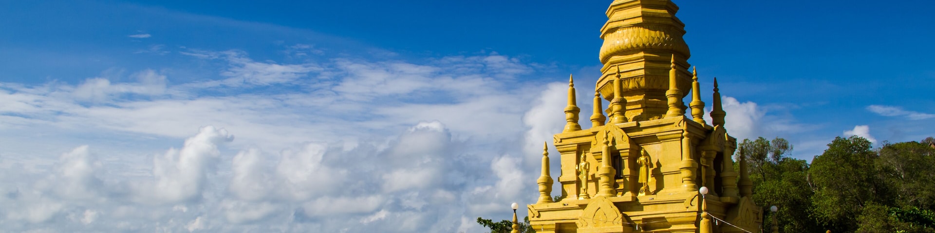 A yellow temple with a pointed spire, situated by a body of water with mountains in the background.