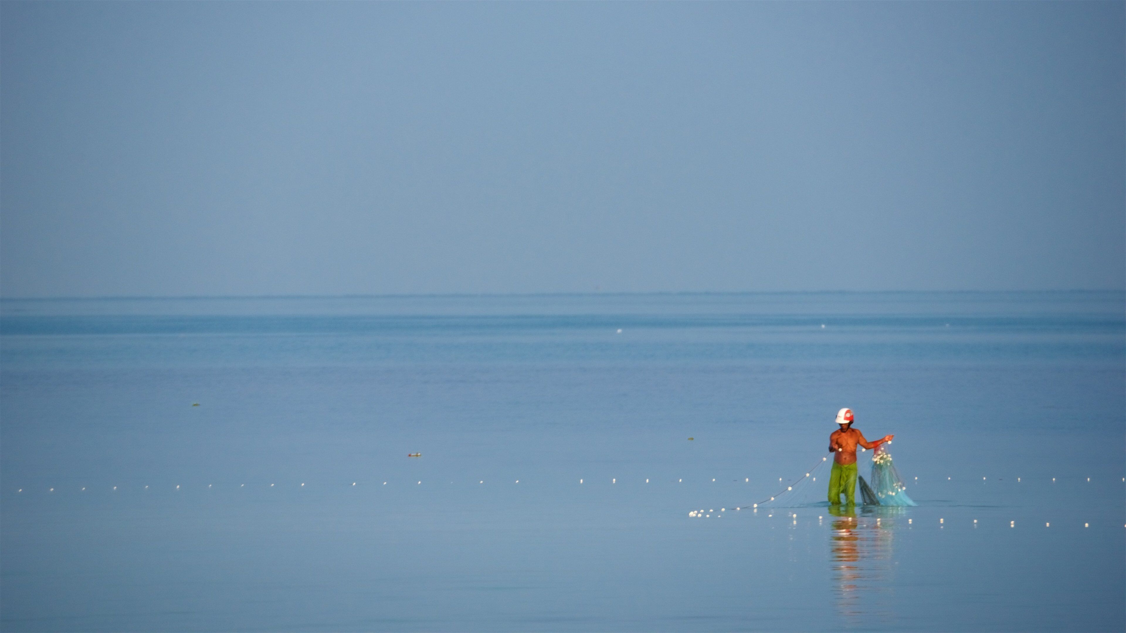 A calm sea with a person in a red cap and green pants standing in shallow water, holding a net.