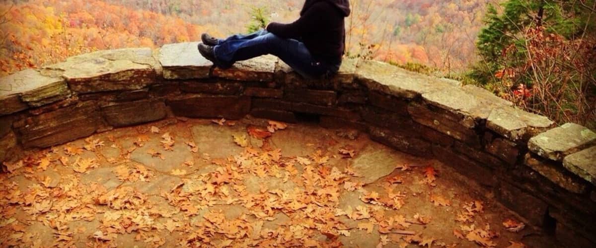 Une personne assise sur un banc de pierre, d’où l’on aperçoit une vallée boisée aux couleurs automnales.