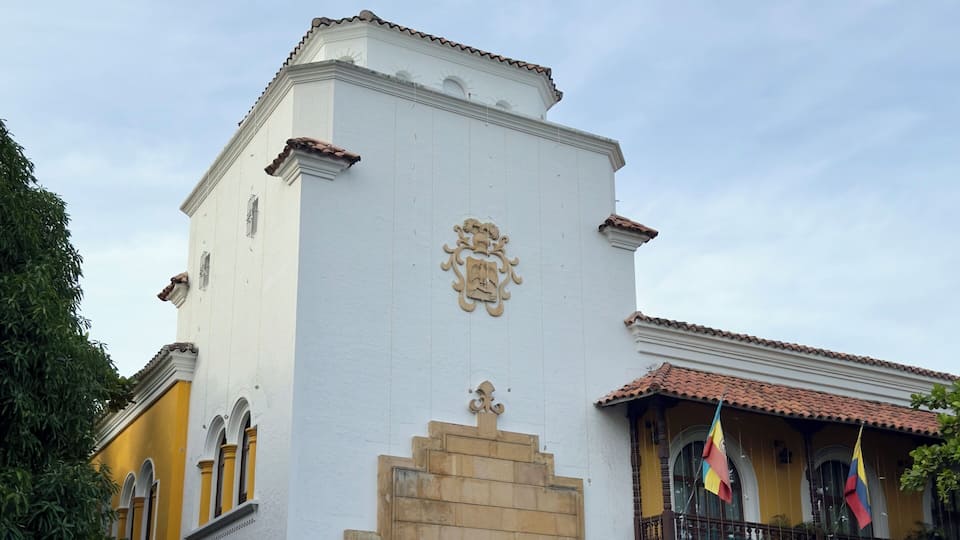 A historic building with a white facade and a yellow section, featuring a decorative emblem and a balcony with flags.