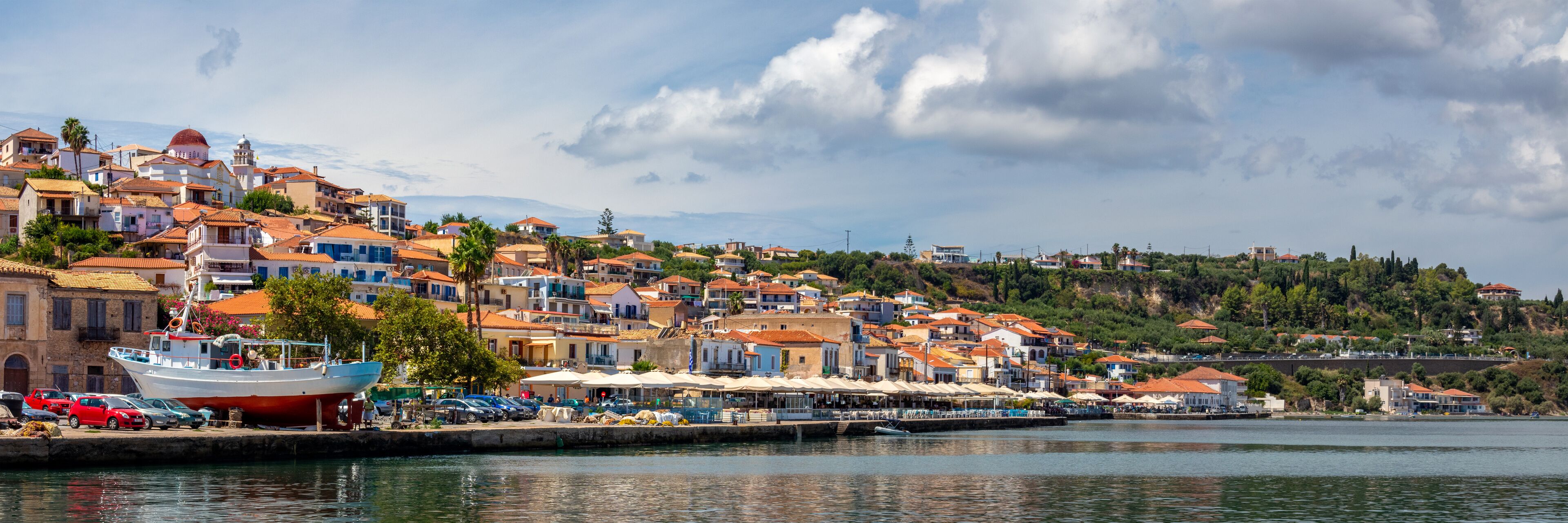 A coastal town with terracotta-roofed buildings, a dock with a boat, and a hillside in the background.