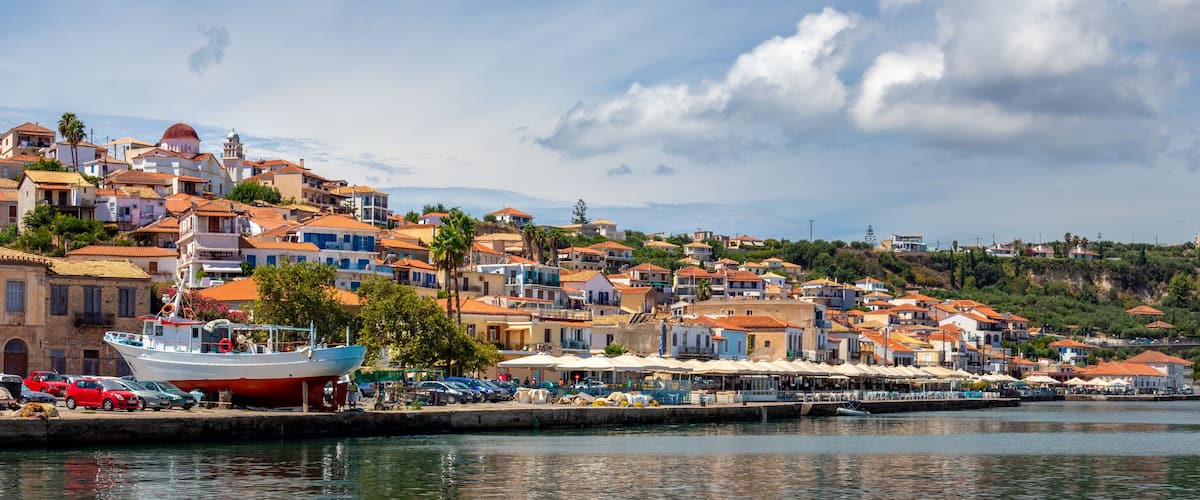 A coastal town with terracotta-roofed buildings, a dock with a boat, and a hillside in the background.