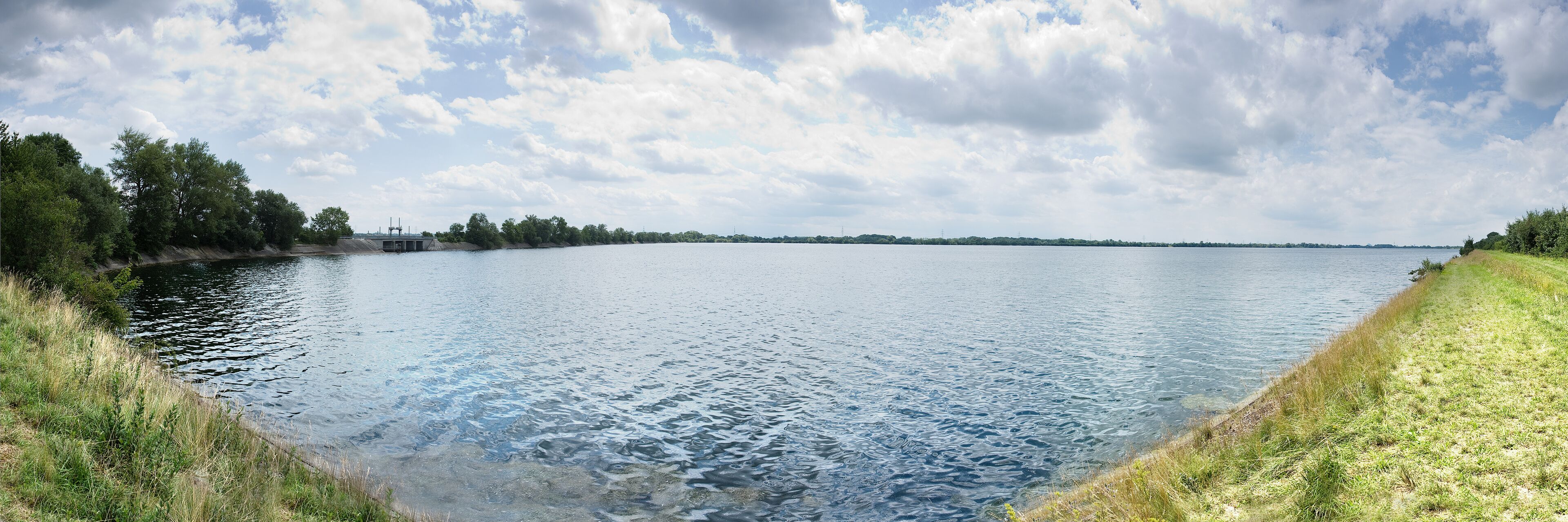 A calm lake with grassy banks, trees, and a cloudy sky.