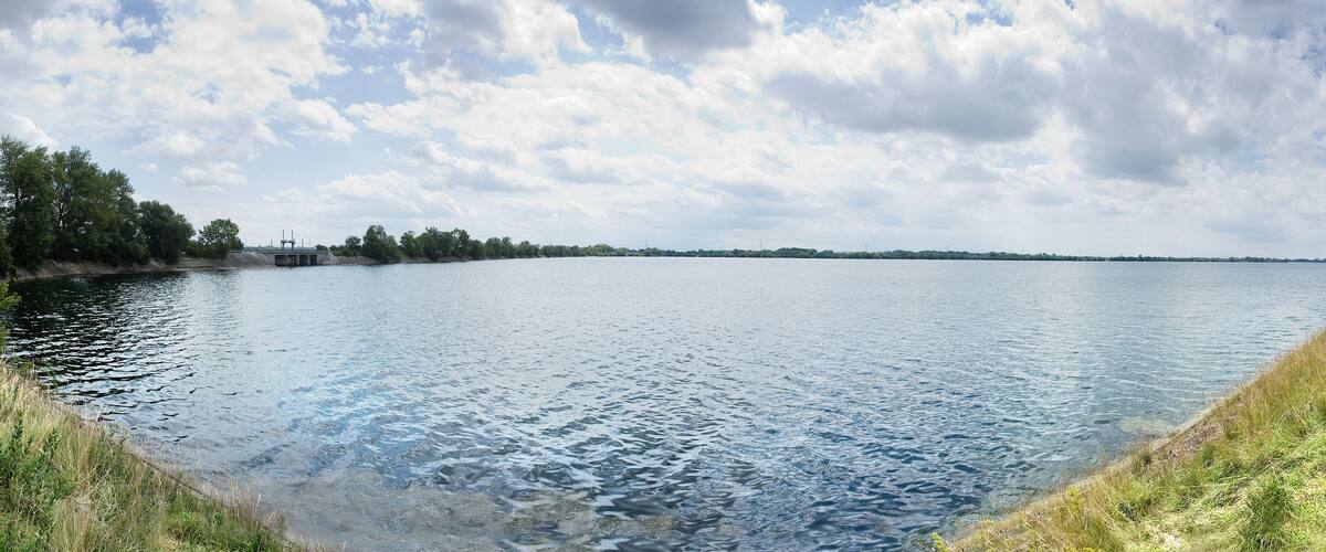 A calm lake with grassy banks, trees, and a cloudy sky.