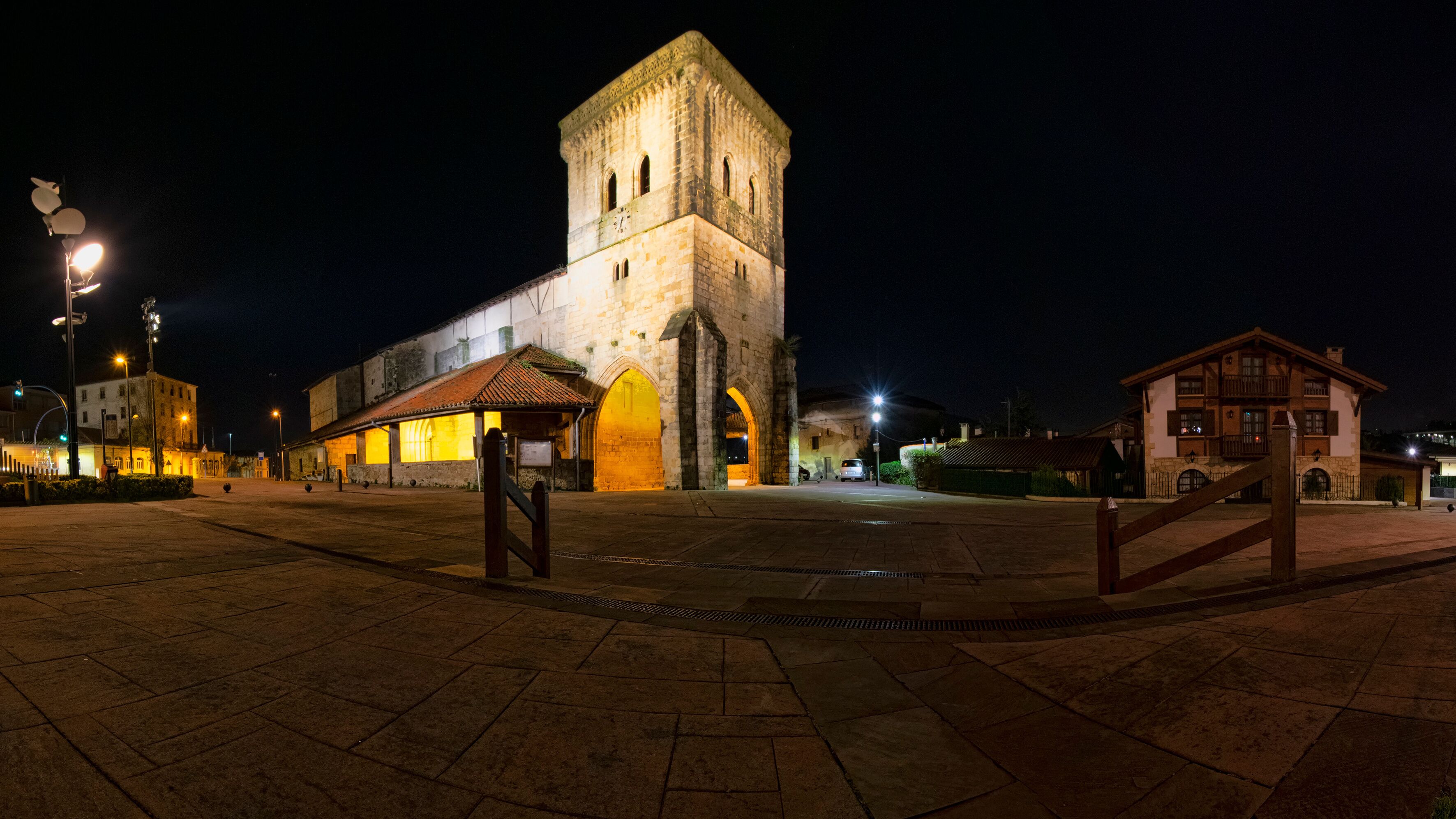 A historic stone tower with arched windows, a plaza with tiled flooring, and a small building with a tiled roof.