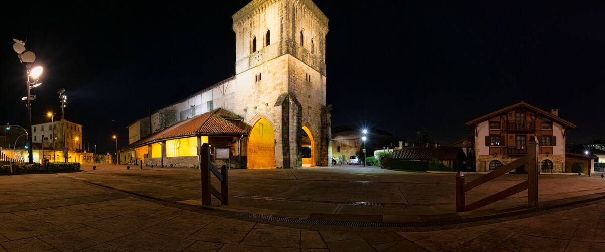 A historic stone tower with arched windows, a plaza with tiled flooring, and a small building with a tiled roof.