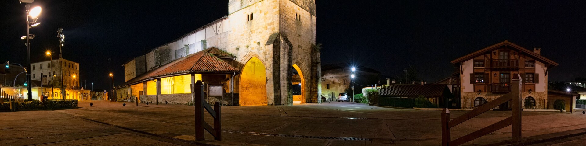 A historic stone tower with arched windows, a plaza with tiled flooring, and a small building with a tiled roof.