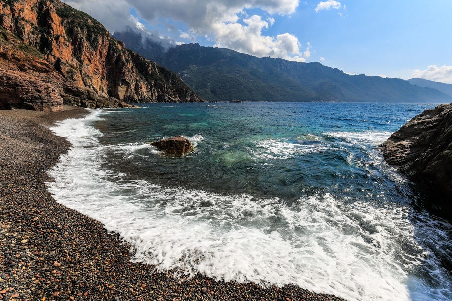 A rocky coastline with clear blue waters and mountains in the background.