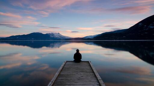 Une personne est assise sur une jetée en bois qui s’avance sur un lac paisible, entourée de montagnes et sous un ciel dégagé.