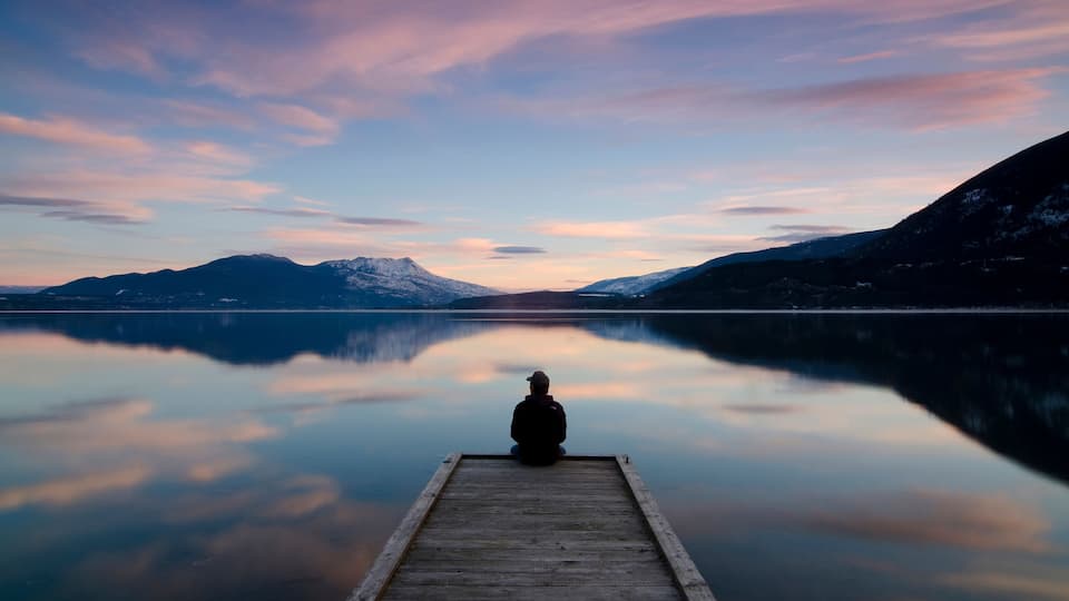 A person sitting on a wooden dock extending into a calm lake, surrounded by mountains and a clear sky.