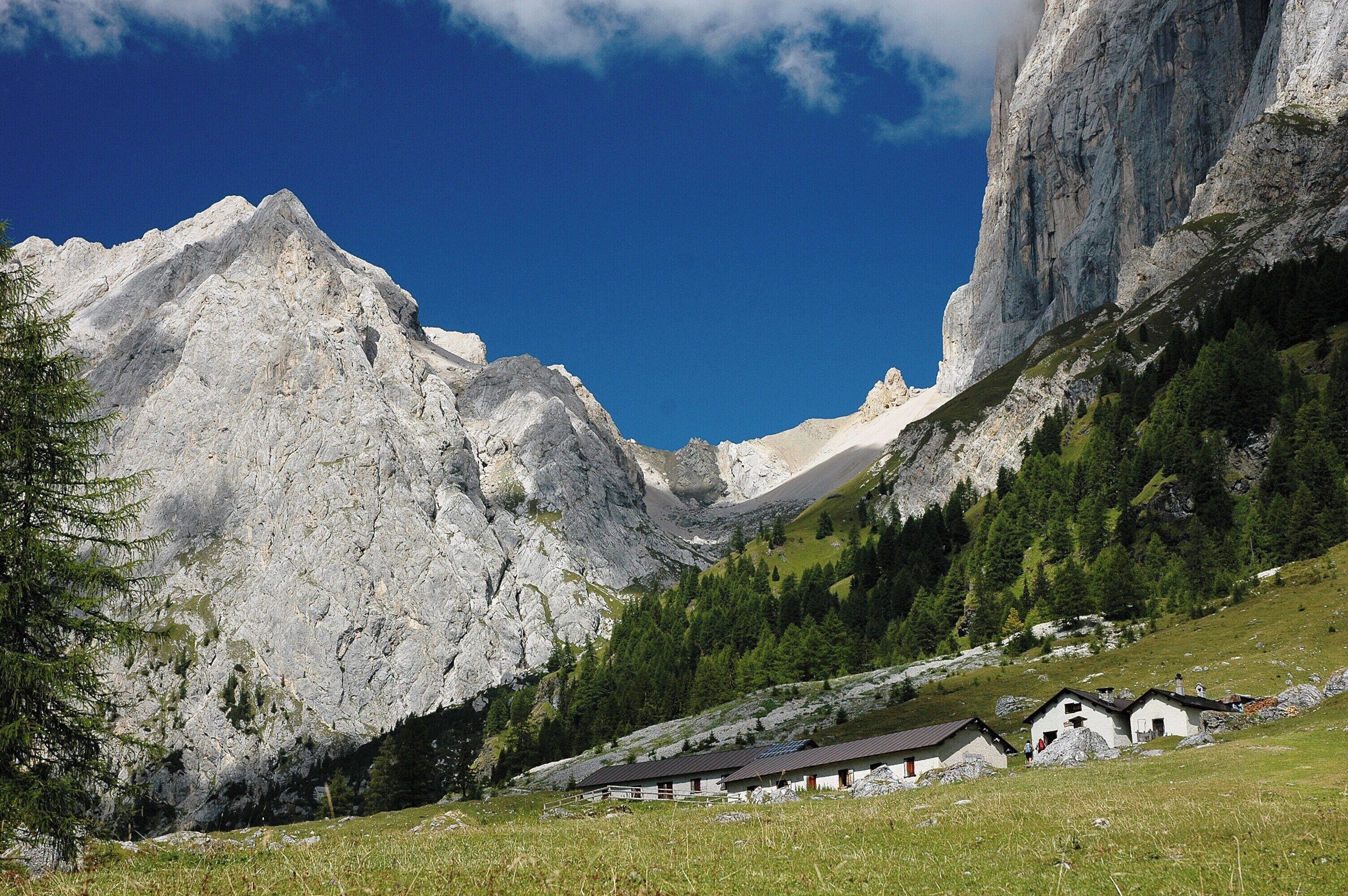 A mountainous landscape with a clear blue sky, rocky peaks, and a small cluster of houses on a grassy slope.
