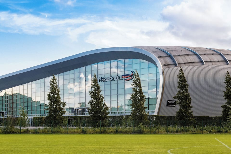 A modern stadium with a curved roof and glass facade, surrounded by a well-maintained lawn.
