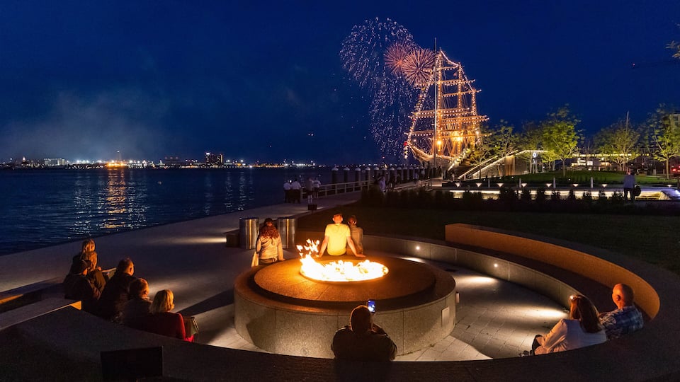 A waterfront with a fire pit, people sitting around it, and a Ferris wheel in the background.