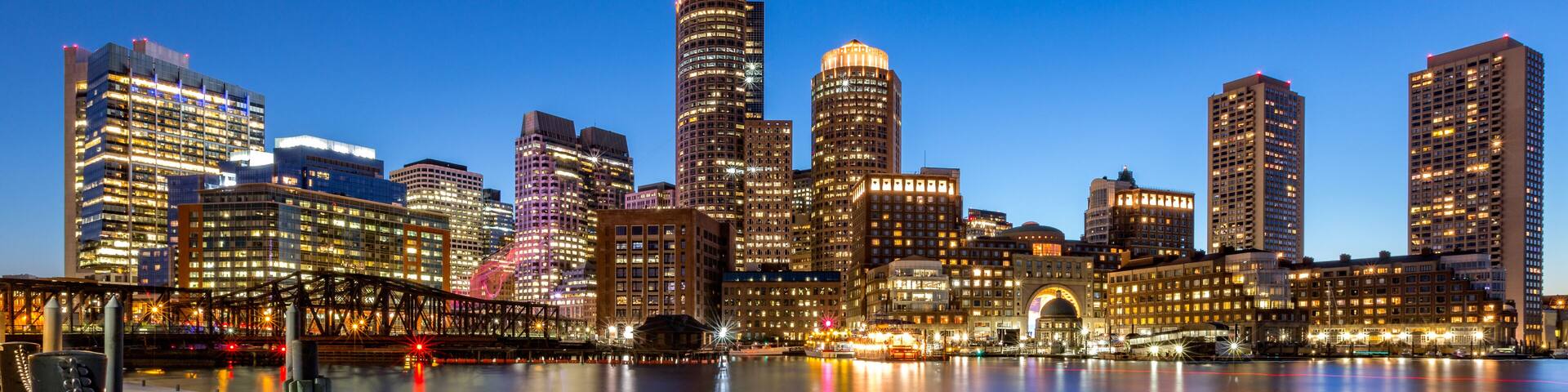 A city skyline at dusk with illuminated buildings reflecting on the water, a prominent bridge, and a large chain barrier along the waterfront.
