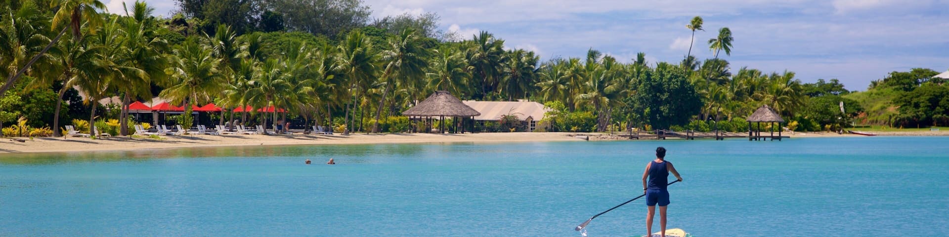 A person paddleboarding on a clear turquoise sea with a beach and palm trees in the background.
