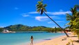 A beach with clear turquoise water, a woman walking, palm trees, and a yacht docked near the shore.