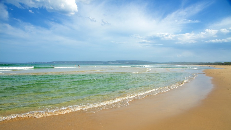 Ein Strand mit klarem türkisblauem Wasser, sanften Wellen und einem weiten Sandstrand unter einer teils bewölkten Himmel.