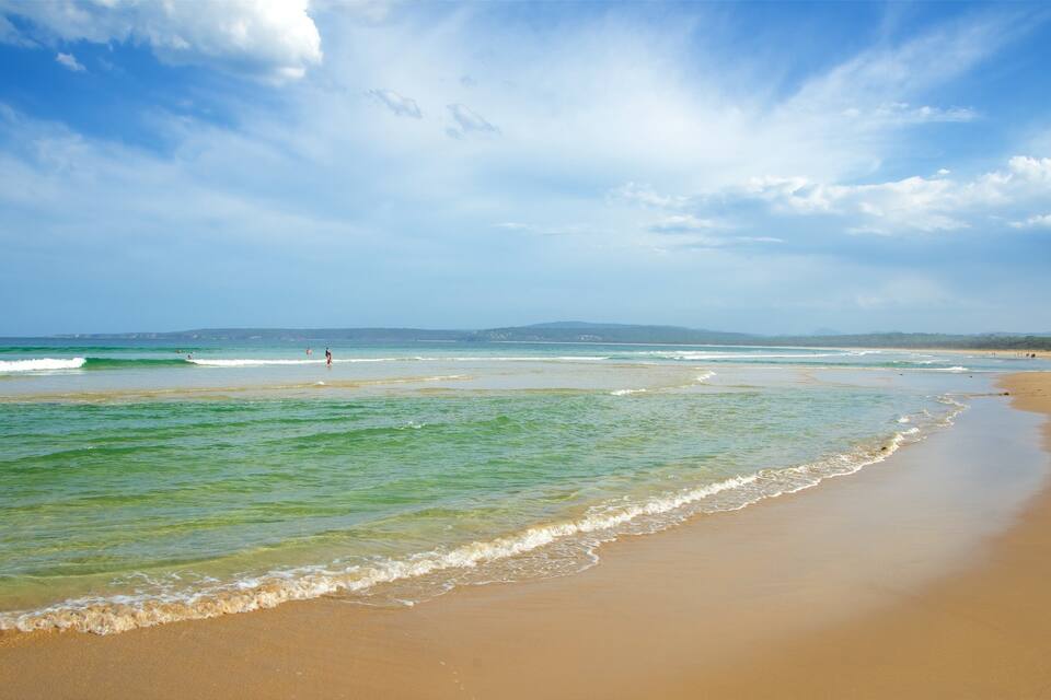 A beach with clear turquoise water, gentle waves, and a wide sandy shore under a partly cloudy sky.