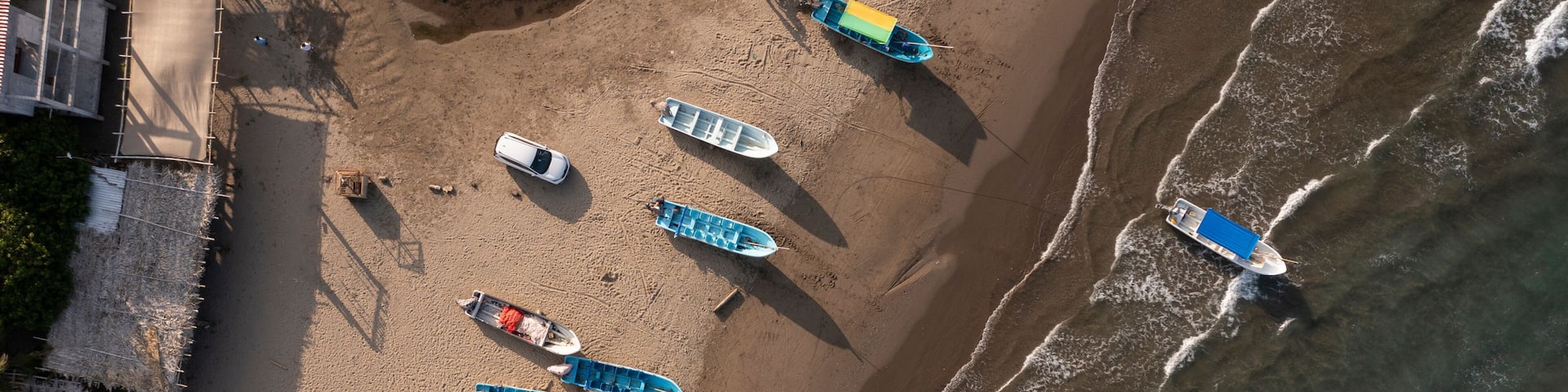 An aerial view of a beach with several boats lined up on the sand, a wooden structure on the left, and waves approaching the shore.