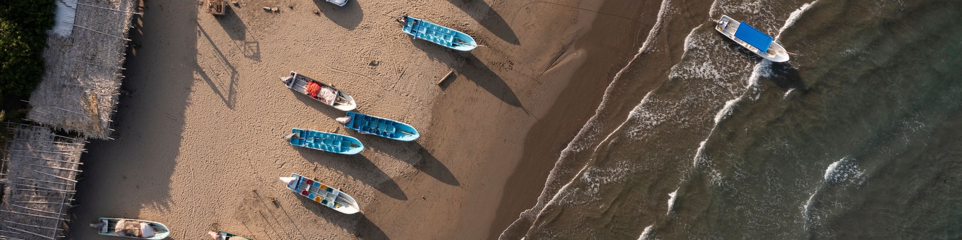 Vista aérea de una playa con varios barcos alineados en la arena, una estructura de madera a la izquierda y olas acercándose a la orilla.