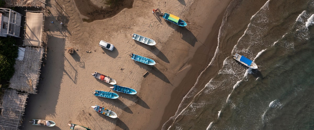 An aerial view of a beach with several boats lined up on the sand, a wooden structure on the left, and waves approaching the shore.