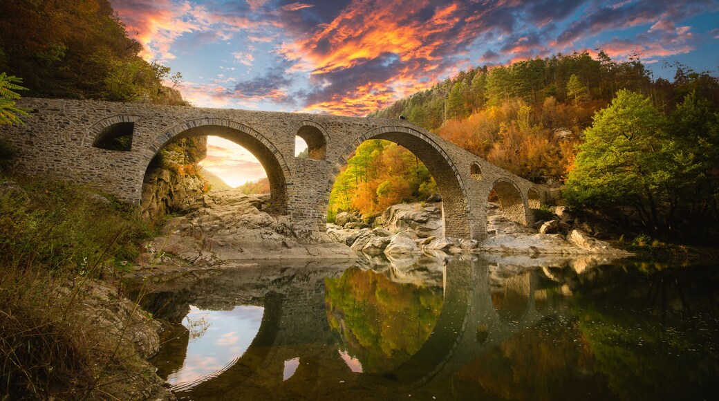 A stone bridge over a river with autumn foliage and a sunset sky.