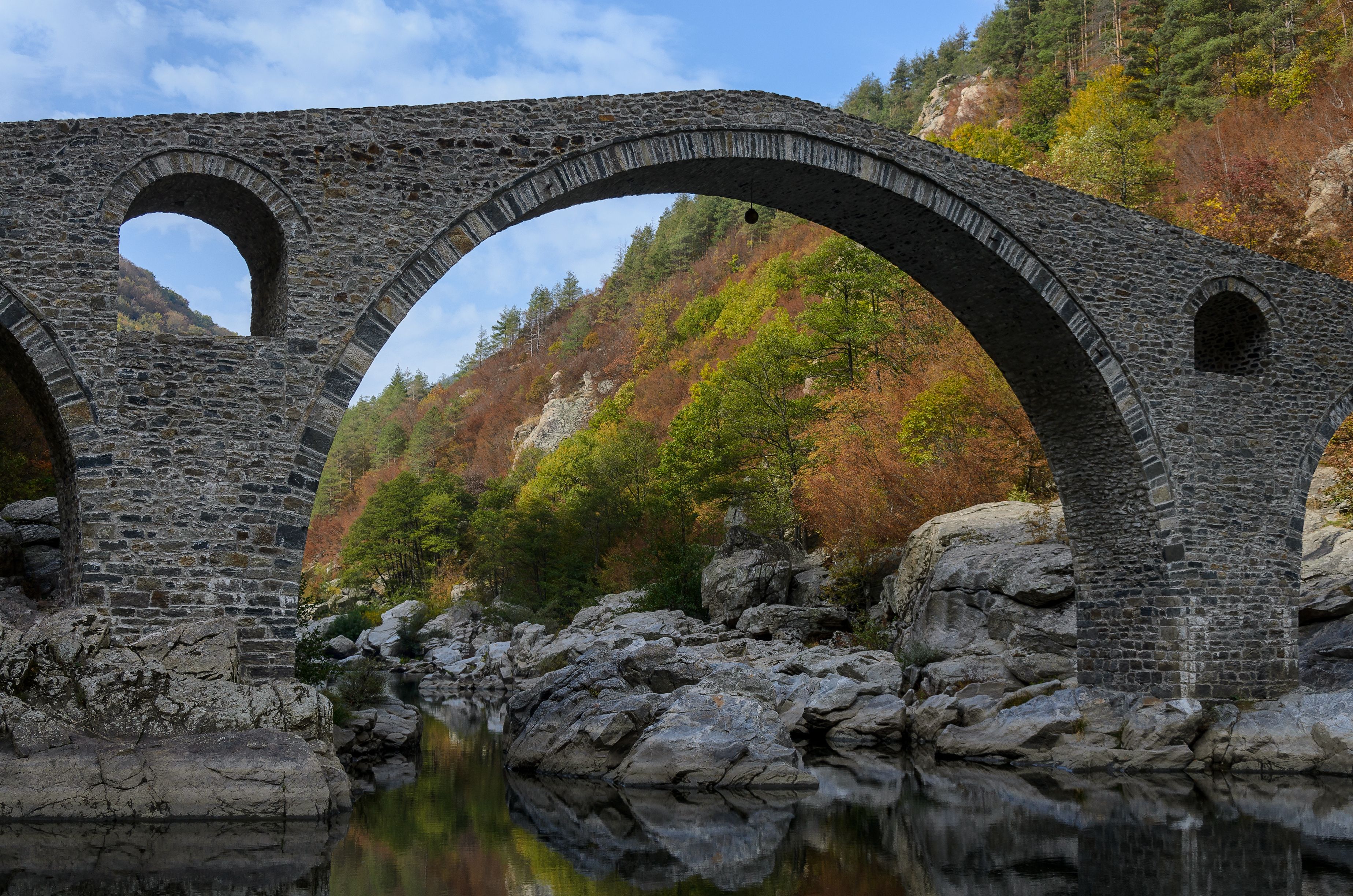 A stone bridge with three arches over a river, surrounded by autumn foliage and rocky terrain.