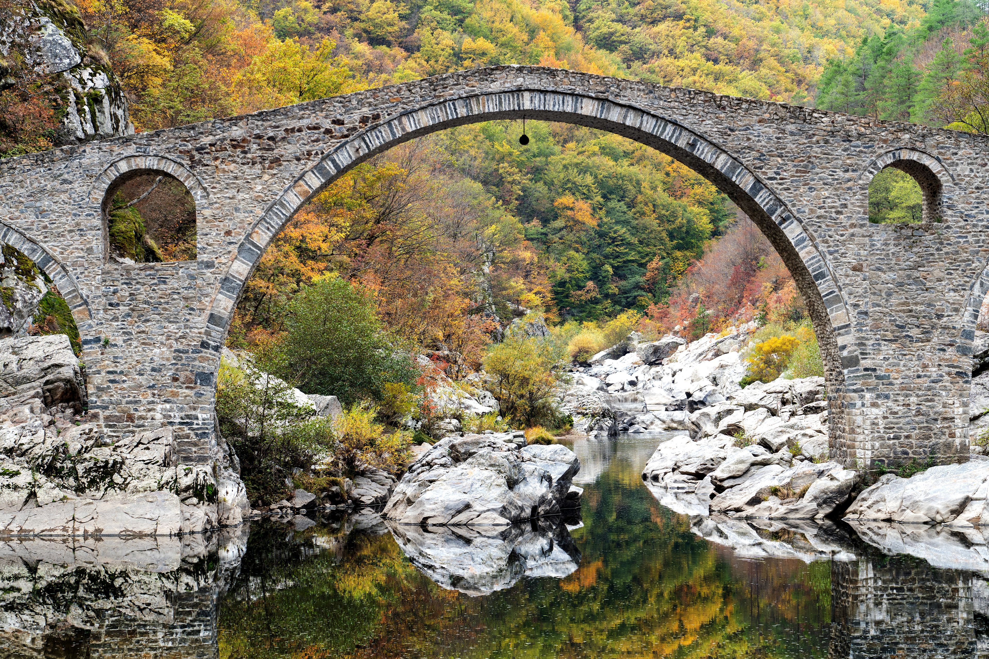A stone bridge arching over a river with autumn foliage in the background.