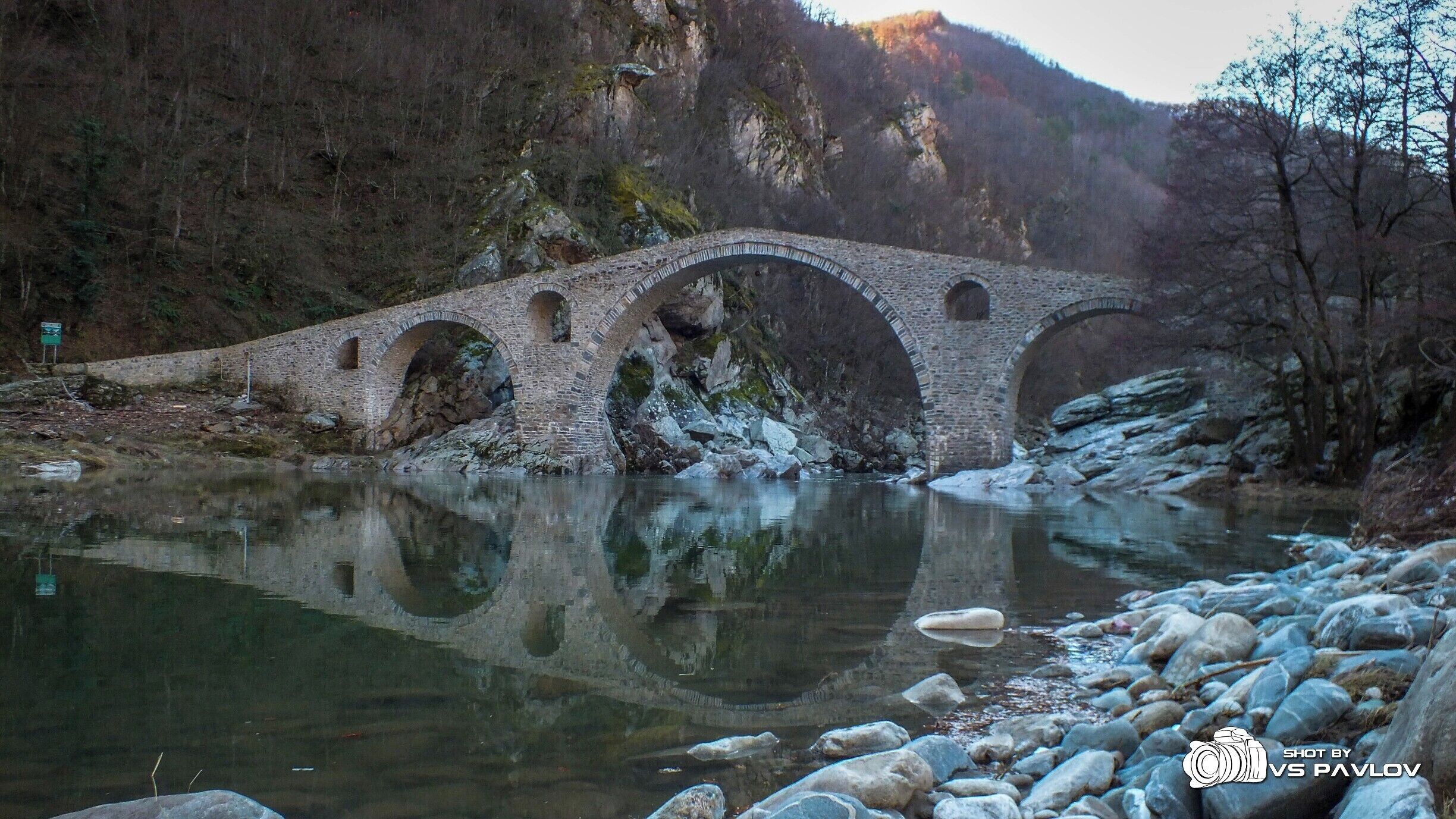 A stone bridge over a river with rocky banks and forested hills in the background.