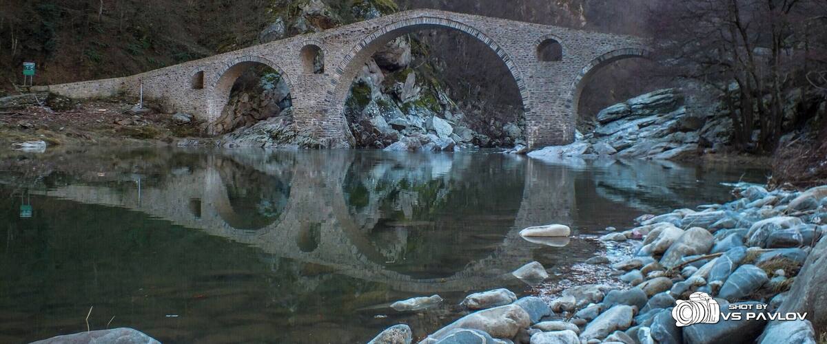 A stone bridge over a river with rocky banks and forested hills in the background.