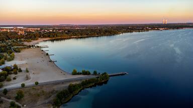 A coastal landscape with a sandy beach, a calm sea, and a distant cityscape with industrial structures.