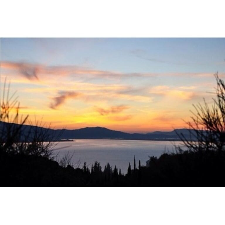 A sunset over a calm lake with silhouettes of trees and distant mountains.