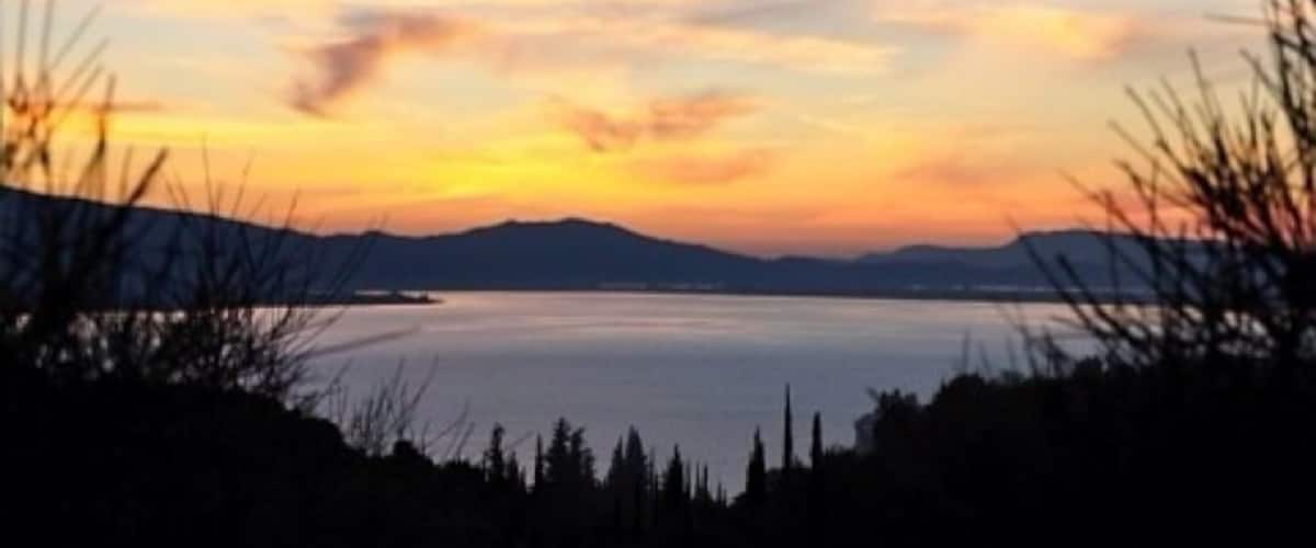 A sunset over a calm lake with silhouettes of trees and distant mountains.