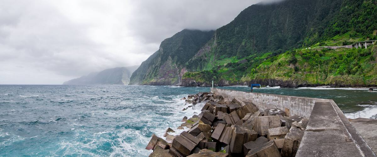 A coastal scene with a rocky breakwater, steep green cliffs, and turbulent ocean waves.