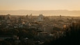 A cityscape at sunset with prominent historical architecture, including domed buildings and a central monument.