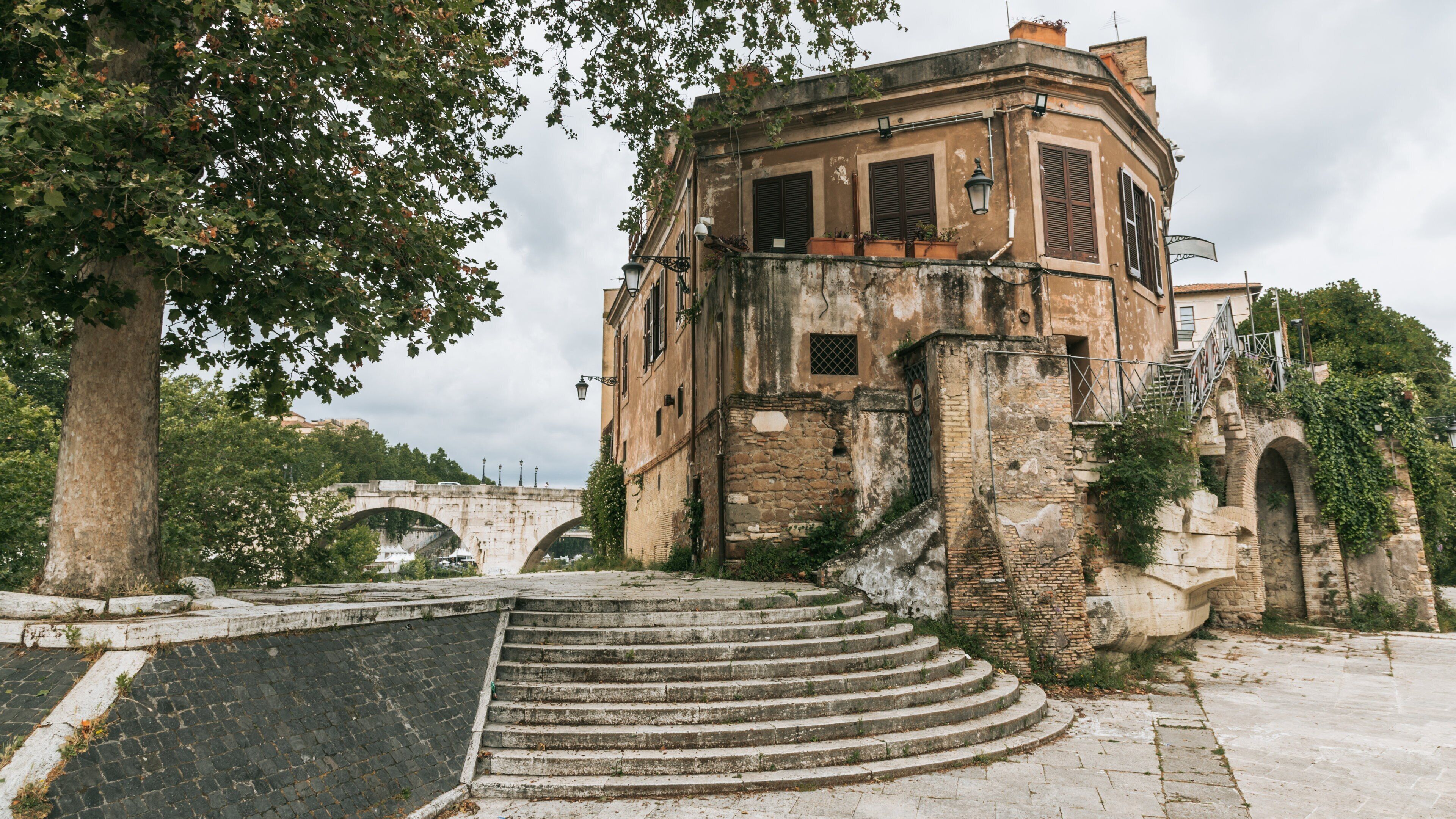 A historic building with a stone staircase leading up to it, surrounded by trees and a stone bridge in the background.
