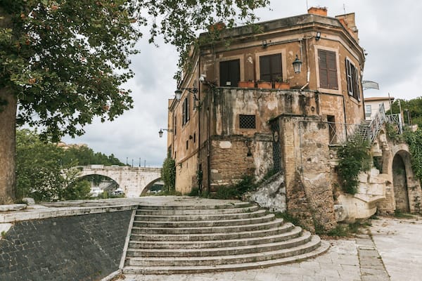 Un bâtiment historique doté d’un escalier en pierre menant à l’entrée, entouré d’arbres et avec un pont de pierre en arrière-plan.