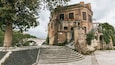 A historic building with a stone staircase leading up to it, surrounded by trees and a stone bridge in the background.