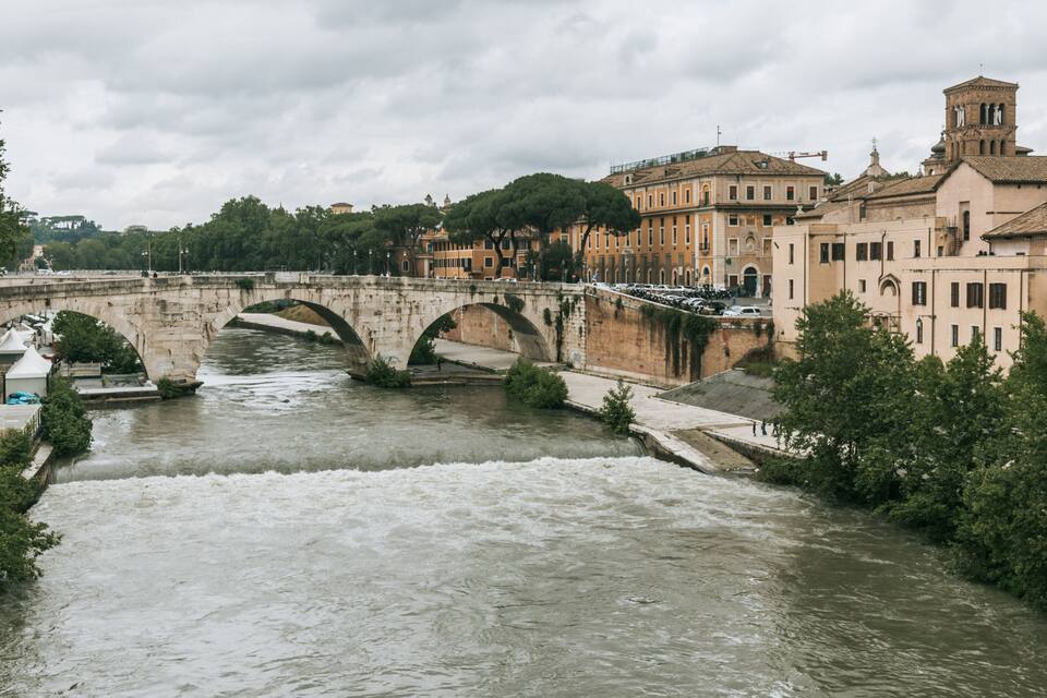 Un riu amb un pont de pedra, edificis històrics i arbres.
