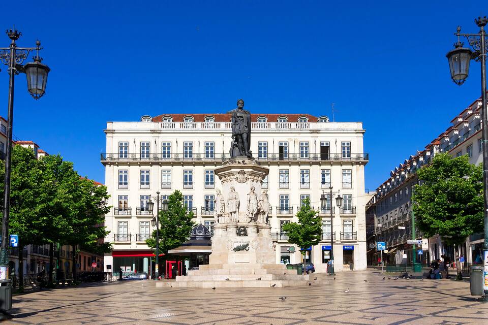 Une place avec un monument central, entourée de bâtiments historiques dotés de balcons et de toits recouverts de tuiles rouges.