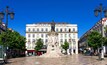 Una piazza con un monumento centrale, circondata da edifici storici con balconi e tetti in tegole rosse.