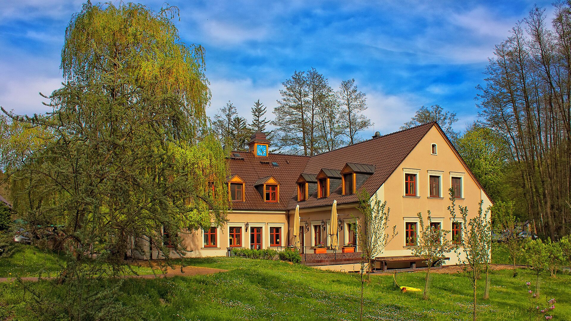 A two-story house with a brown roof and yellow walls, surrounded by trees and a grassy lawn.